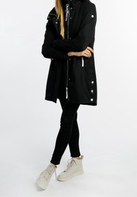 Personne portant une veste noire avec des fermetures éclair et des boutons blancs, un pantalon noir et des baskets montantes beige clair, debout les bras croisés.