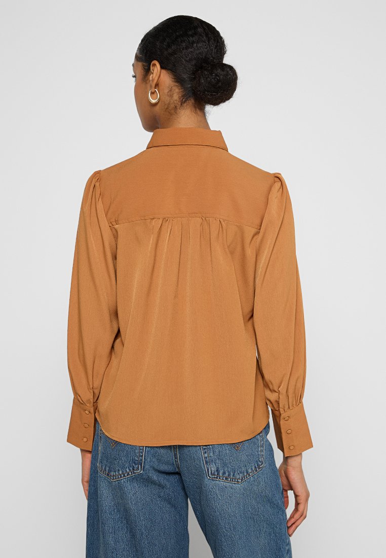 Vrouw met donker haar in een lage knot, gekleed in gouden oorbellen, een bruin blouse met lange mouwen en knoopmanchetten, en blauwe spijkerbroek, met de rug naar de kijker gekeerd.