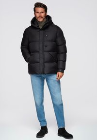 Chaqueta negra acolchada con capucha, que presenta amplias secciones acolchadas, un collar alto y bolsillos laterales. Lleva puesta con jeans de mezclilla azul claros y botas negras.