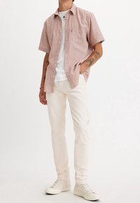 Vaaleanpunainen lyhythihainen nappipaita valkoisen t-paidan päällä, yhdistetty vaaleanbeigeihin farkkuihin ja valkoisiin tennareihin, käsivarsissa näkyvillä tatuointeja.