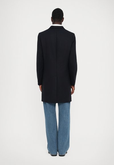 Cappotto in misto lana blu navy con silhouette strutturata, spacco singolo e vestibilità su misura, abbinato a jeans in denim azzurro a gamba larga.