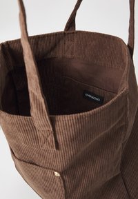 Barna kordbársony tote táska függőleges bordázással, két vállpánttal és egy kisebb belső zsebbel, amelyen fekete címke található.