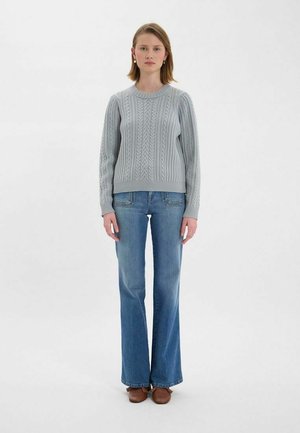 Pull en tricot gris à texture côtelée, avec un col rond et un motif en câble. Associé à un jean bleu clair évasé et des chaussures marron.