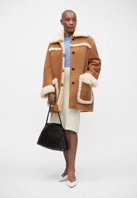 Cappotto in shearling marrone con dettagli bianchi, bottoni e grandi tasche. Top azzurro chiaro e gonna bianca. Borsa a tracolla nera intrecciata e scarpe color crema.