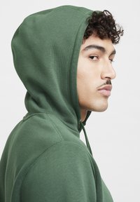 Felpa con cappuccio verde realizzata in tessuto morbido, con un design aderente, cordino e dettagli di cucitura a vista. Texture liscia.