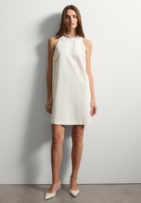 Vestido blanco sin mangas con un escote alto, tejido suave y un sutil detalle de cuello. Combinado con zapatos de tacón beige puntiagudos.