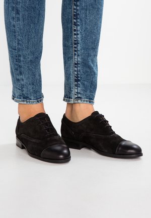Black suede shoes with a smooth leather toe cap, lace-up design, and a low heel, paired with blue denim jeans.