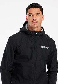 Czarna wodoodporna kurtka z fakturowanym wykończeniem, z pełnym zamkiem błyskawicznym, regulowanym kapturem i widocznym logo na klatce piersiowej. Posiada boczne kieszenie.