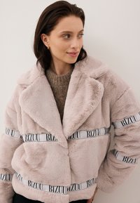 Kurtka z futerka w jasnoróżowym kolorze z szerokimi klapami, z czarno-białymi akcentami w postaci wstążek z logo wokół talii. Strukturalny, puszysty materiał.