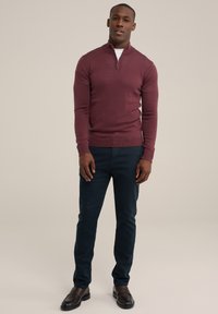 Bordo zip-pletena veste s dugim rukavima u kombinaciji s tamnoplavim hlačama. Glatka tekstura, kroj koji se prilagođava tijelu i okrugli izrez. Braon kožne cipele.