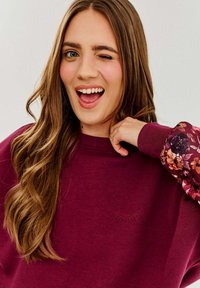Burgundowa bluza z kwiatowymi rękawami, teksturowany materiał i subtelny haft w kształcie rzęs na klatce piersiowej.