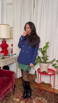 Γυναίκα με μπλε σακάκι και φούστα πράσινη καρό, κρατώντας τσάντα με λεοπάρ σχέδιο, στέκεται εσωτερικά κοντά σε κόκκινο τραπέζι με φυτό και κόκκινο φωτιστικό σε λευκό γραφείο.