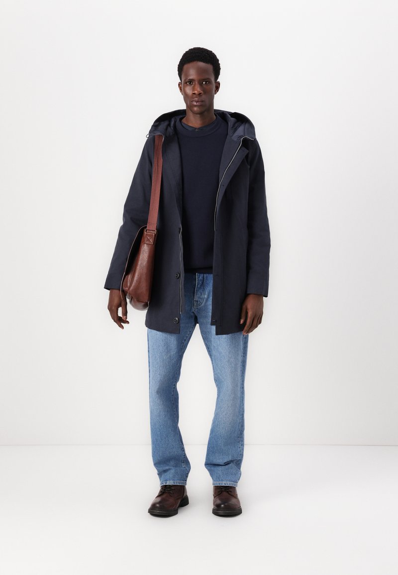 Granatowa wodoodporna kurtka z kapturem, zapinana na guziki, oraz brązowa skórzana torba listonoszka. Jasnoniebieskie dżinsy i ciemne buty dopełniają strój.