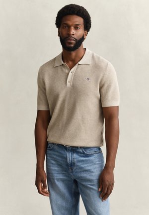 Man met baard, gekleed in een beige korte mouw polo en blauwe spijkerbroek, tegen een effen lichte achtergrond.