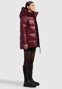 Burgundy puffer jacket with a hood, quilted design, side pockets, and drawstrings. Worn over a black mini skirt and thigh-high black boots.