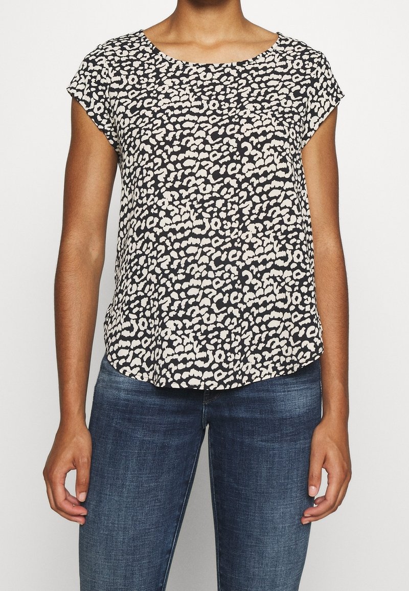 Haut noir et crème à manches courtes avec un motif à pois, décolleté rond et coupe ample, porté avec un jean bleu.