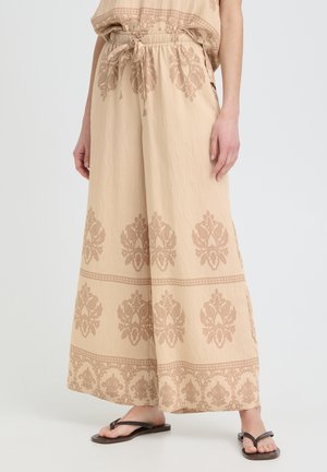 Pantaloni beige a gamba larga con coulisse in vita e motivi floreali marroni, indossati con infradito nere su sfondo semplice.