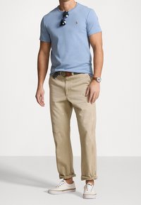 Muž v světle modré košili s krátkým rukávem, béžových nohavicích vyhrnutých nahoru s barvami, bílých nazouvacích botách, hodinkách a slunečních brýlích zavěšených na košili.