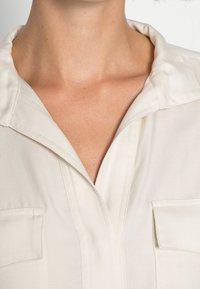 Weißes Hemd mit Kragen aus strukturiertem Stoff, das ein subtil diagonales Muster aufweist und auf beiden Seiten aufgesetzte Taschen hat.