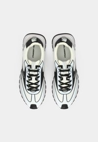 Ténis desportivos brancos e pretos com parte superior lisa em material sintético, atacadores proeminentes e sola de borracha texturada. Apresentam biqueira arredondada e logótipo.