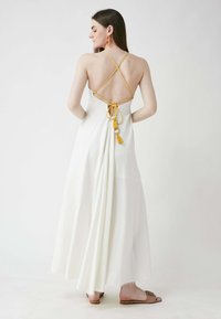 Weißes Maxikleid mit überkreuzten Rückenträgern. Gelbe Akzente und Quasten verzieren die Taille. Der glatte Stoff fließt bis zum Boden.