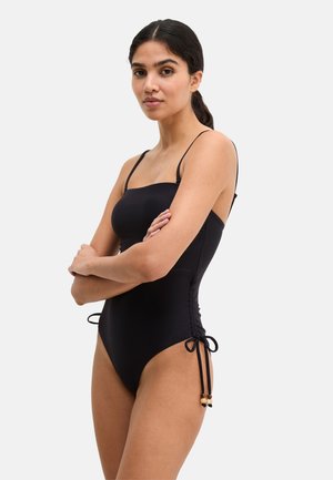 Femme aux cheveux foncés portant un maillot de bain une pièce noir avec fines bretelles et liens latéraux, debout bras croisés devant un fond uni.