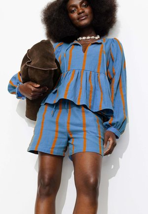 Femme aux grands cheveux bouclés portant un haut à manches longues rayé bleu et orange avec un short assorti, tenant une veste en daim marron pliée.