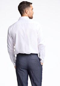 Camicia bianca a maniche lunghe con colletto classico, abbinata a pantaloni blu scuro con due tasche posteriori e un tessuto texturizzato.