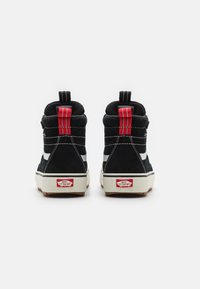 Svart semsket høyhælte sneakers med røde hælaksenter og hvite striper. Har en hvit gummisåle og et merket rødt etikett på hælen.