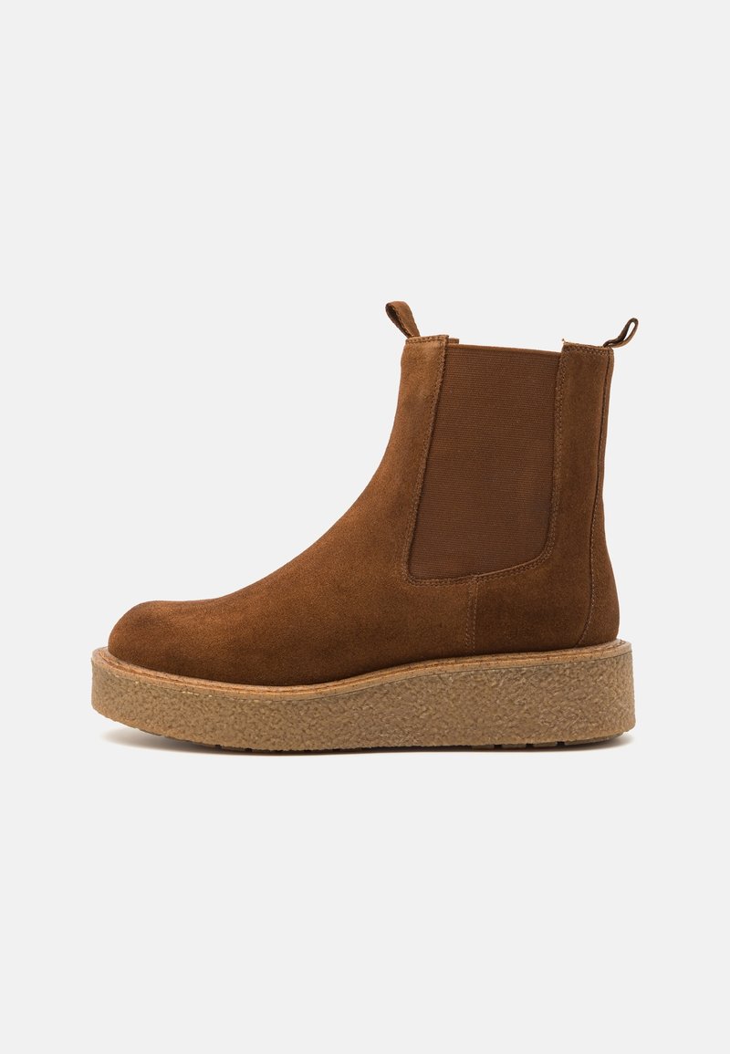 Botte Chelsea en daim marron avec une semelle épaisse texturée, des panneaux élastiques sur les côtés et des languettes de traction à l'avant et à l'arrière sur un fond blanc uni.