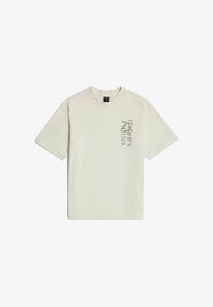 Beżowy T-shirt z krótkim rękawem, okrągłym dekoltem i małym pionowym napisem na lewej stronie klatki piersiowej.