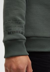 Green sweatshirt with ribbed cuffs, featuring textured black print details on the sleeve. Soft fabric and relaxed fit.
