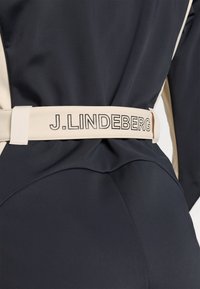 Melnas kombinezons ar bēšiem akcentiem un platām jostām, uz kuras melnā burtnīcā ir rakstīts "J. LINDEBERG". Audums izskatās gluds un pieguļošs.