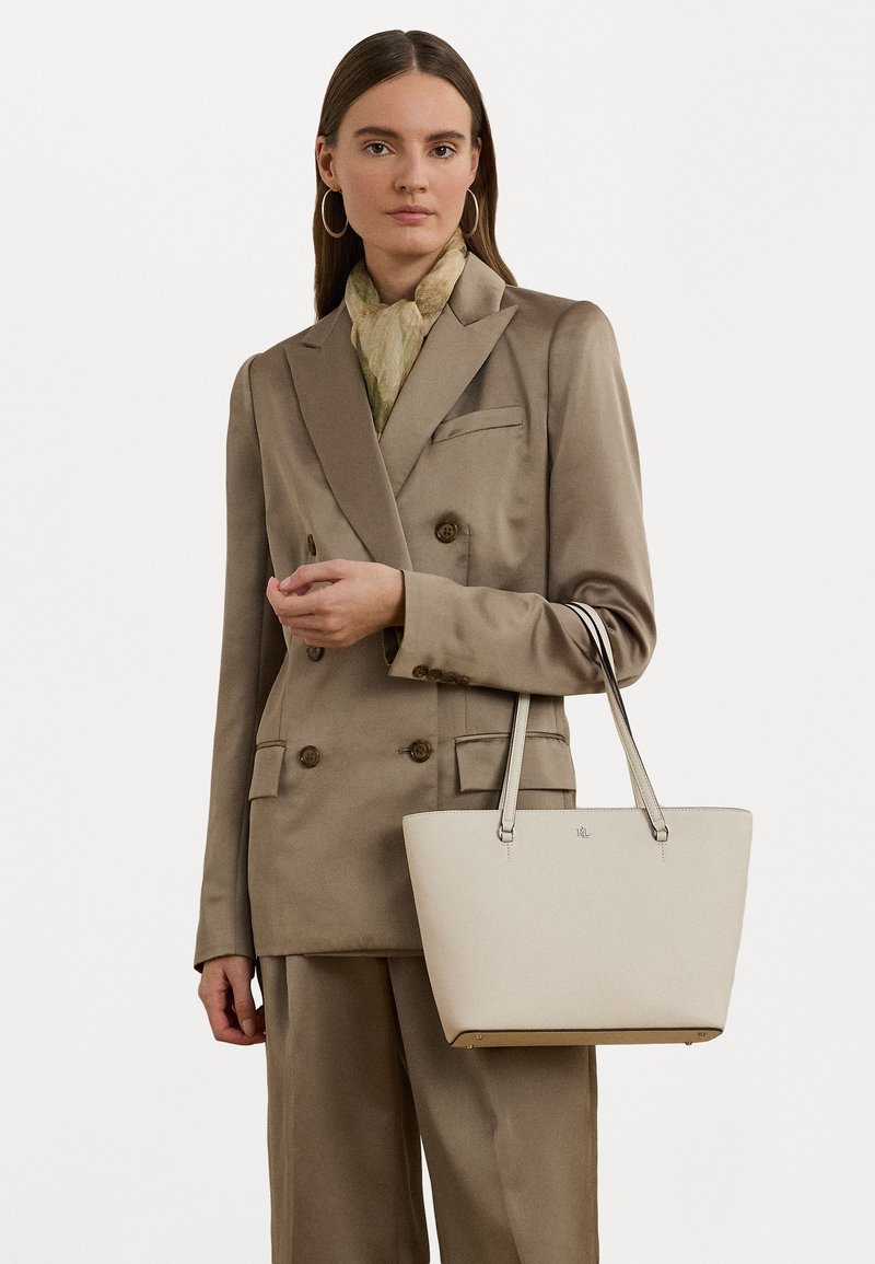 Bolsa tote bege em couro suave, com detalhes em metal prateado e alças de transporte duplas, combinada com um fato taupe e um lenço claro.