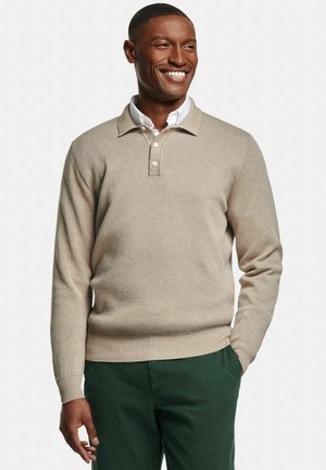 Homme souriant portant un pull beige boutonné sur une chemise blanche et un pantalon vert, debout avec une main dans la poche devant un fond uni.
