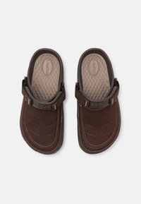 Sandales en cuir marron avec une semelle en caoutchouc texturé et une sangle réglable, présentant une surface supérieure lisse avec des accents de couture.