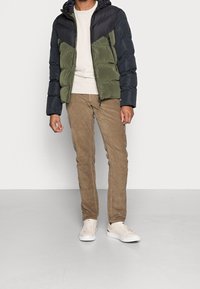 Puffjakke i sort og olivengrøn, med lynlås foran, hætte og quiltet design. Sammen med tan farvede kordfløjlsbukser og beige sneakers.