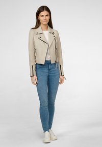 Cremfarbener Lederbikerjacke mit schwarzen Reißverschlussakzenten, langen Ärmeln und seitlichen Reißverschlusstaschen, kombiniert mit enganliegenden blauen Jeans und weißen Sneakern.