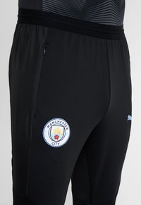 Primer plano de pantalones deportivos negros con el escudo del Manchester City en el muslo izquierdo y el logo de Puma en azul claro en el muslo derecho.