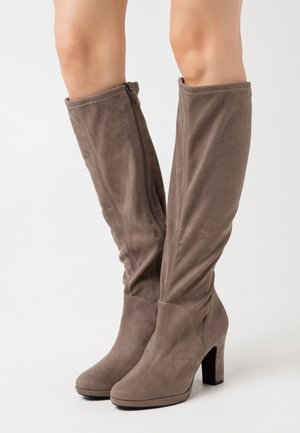 Bottes montantes en daim marron avec fermeture éclair latérale et talons blocs portées sur jambes nues sur fond blanc.