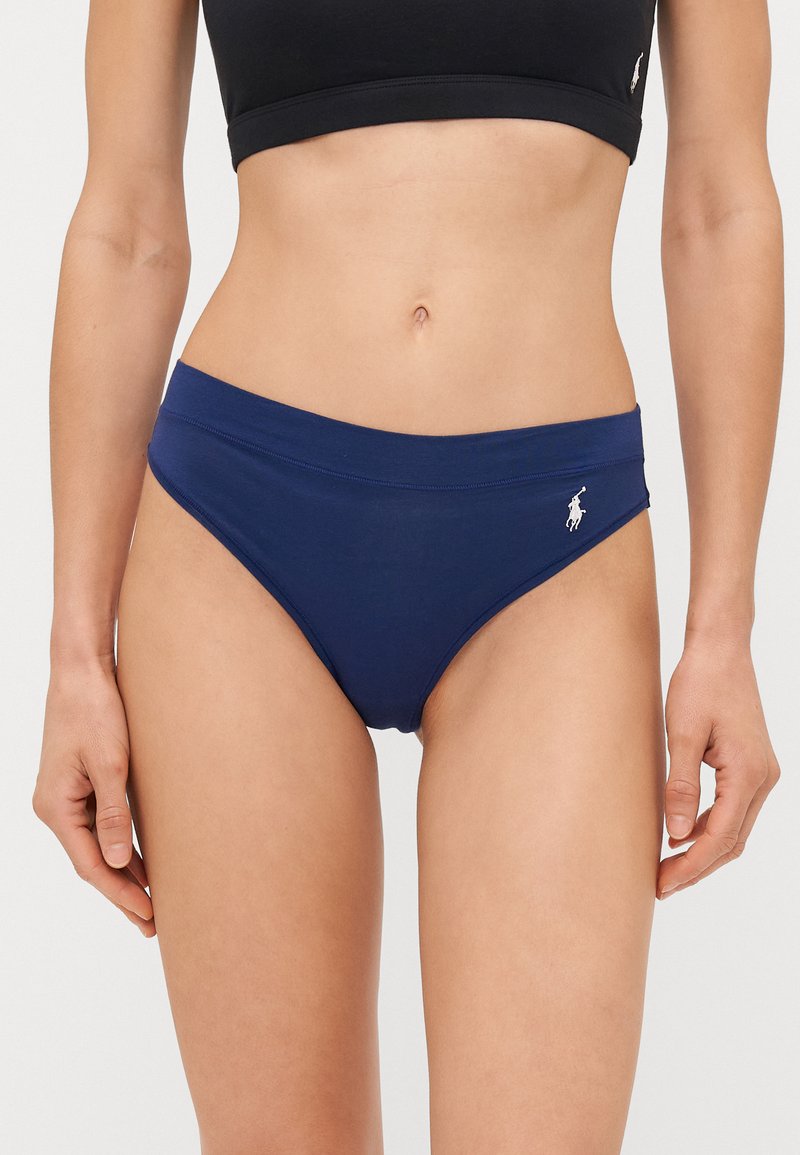 Tumša zilā bikini apakšdaļa ar gludu tekstūru, piedāvā plašu jostasvietu un nelielu baltu logotipu uz sāna.