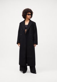 Lang sort frakke lavet af tykkere materiale, med en dobbeltbrystet design, store knapper, tilpassede skuldersømme og sidelommer.