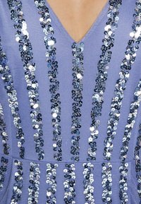 Lila šaty svisle pruhované třpytivými modrými a stříbrnými flitry. Výstřih do V a texturovaná látka.