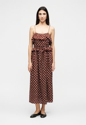 Žena s dlhými tmavými vlasmi, oblečená v hnedých midi šatách s bielymi bodkami, tenkými ramienkami, volánmi a čiernymi balerínami, stojaca pred jednofarebným pozadím.