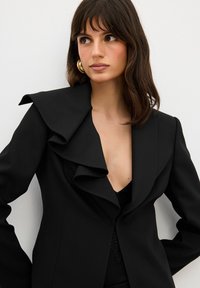 Blazer nero con colletto arricciato, spalle strutturate e scollatura profonda a V. Tessuto liscio, design aderente e stile minimalista.