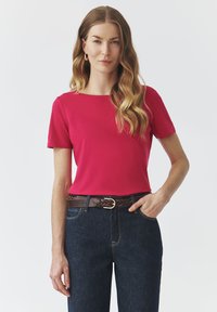 Un haut rose vif à manches courtes avec un col rond, associé à un jean en denim bleu foncé et une ceinture en cuir tressé marron.