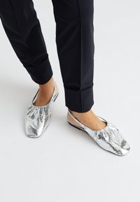 Zilveren metallic platte schoenen met een verzameld ontwerp en open achterkant, met een vierkante neus en een gladde textuur, gecombineerd met zwarte broeken.
