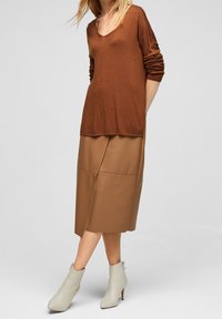 Haut marron en maille à manches longues avec un col en V, associé à une jupe midi en cuir beige avec des coutures latérales et une fente. Bottines blanches à la cheville.