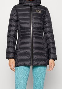 Svart vadderad jacka med huva, med dragkedja, horisontell sömnad och en märkeslogotyp på bröstet. Slätt, glänsande material.