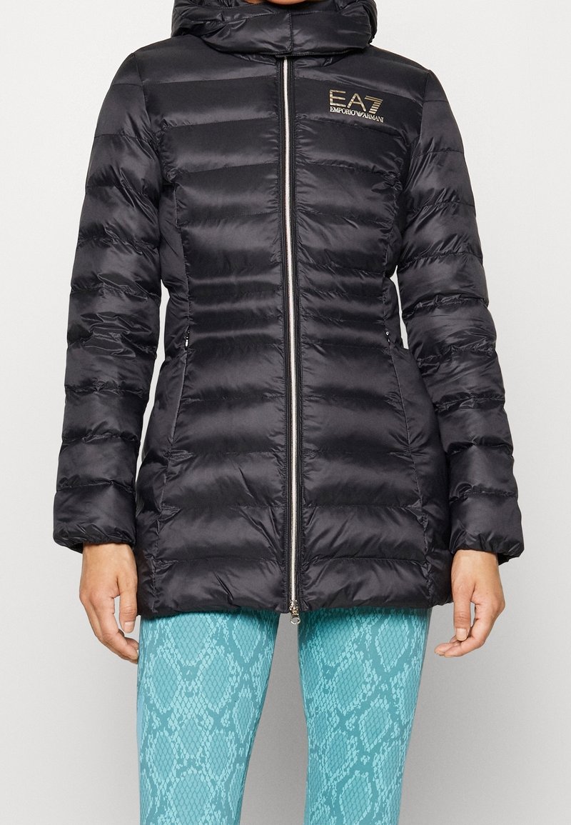 Svart vadderad jacka med huva, med dragkedja, horisontell sömnad och en märkeslogotyp på bröstet. Slätt, glänsande material.